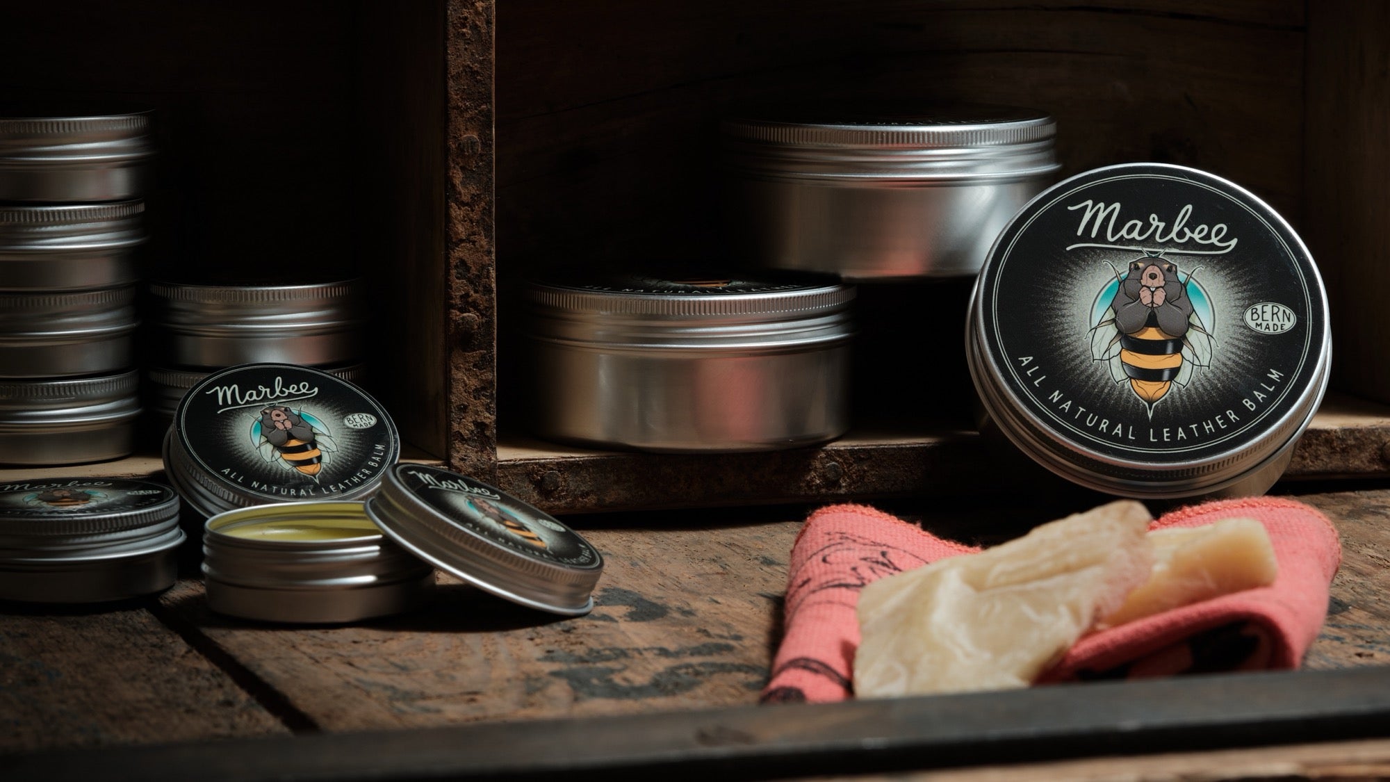 Marbee All Natural Leather Balm von oldpassion: offene Dose mit Balsam, Pflegetuch und Bienenwachs auf einer warmen Holzkiste, Thema Ledergürtel pflegen.
