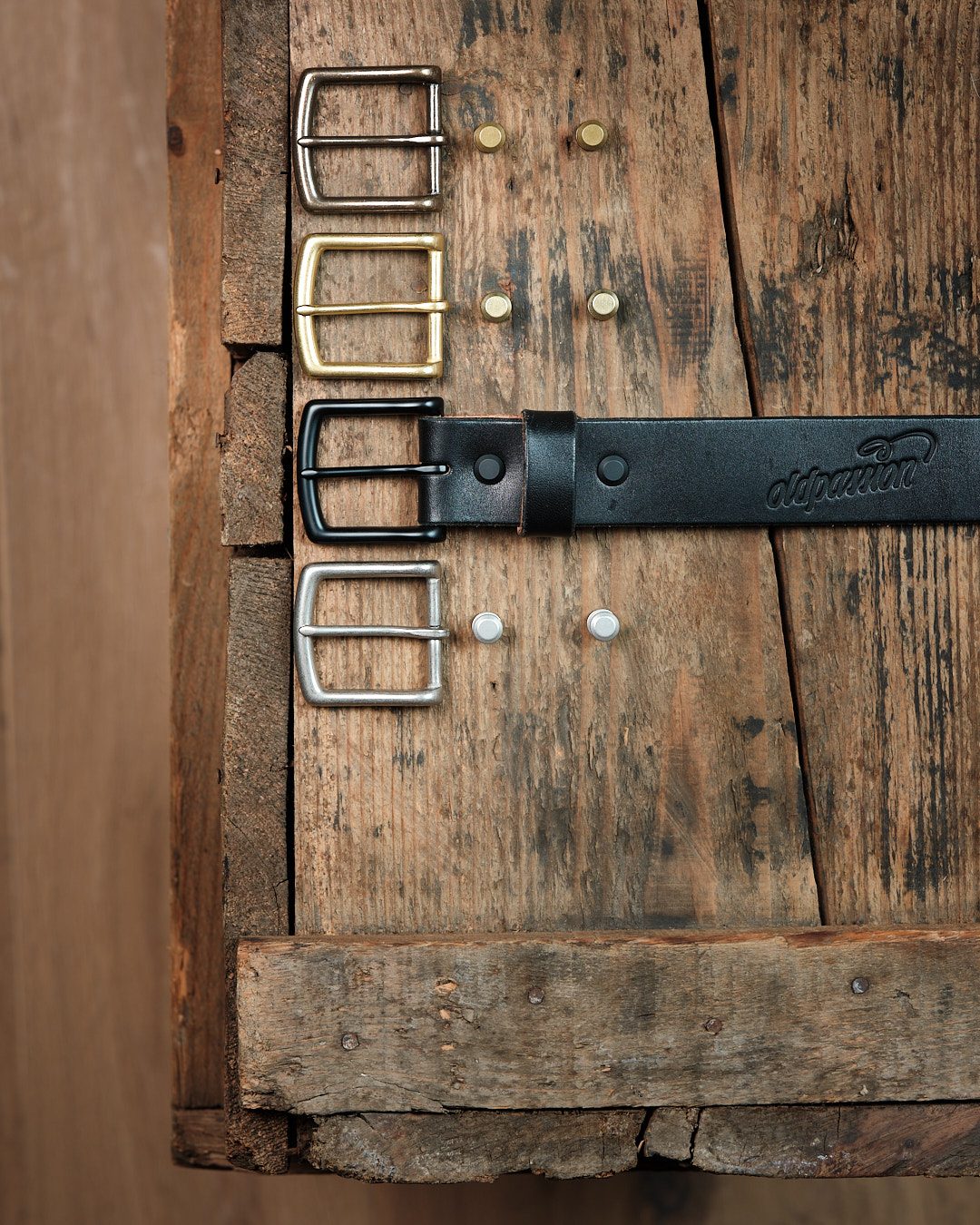 Schwarzer oldpassion Ledergürtel mit mehreren wechselbaren Messing- und Stahlschnallen auf rustikalem Holz, Thema Lederwaren als Geschenk.